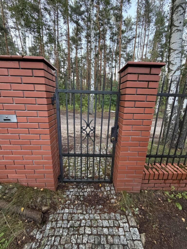 Czarna furtka pomiędzy ceglanymi słupkami, z prętami i ozdobnym elementem w środku.